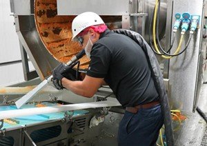 Técnico con equipo de seguridad realizando una limpieza industrial en maquinaria de una planta de alimentos y bebidas para eliminar residuos sin usar productos químicos.